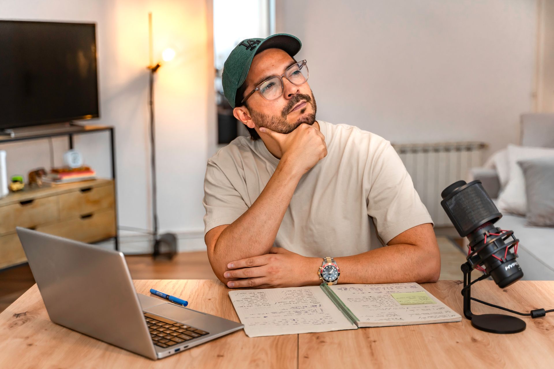 Un hombre con gorra, gafas y reloj contempla, sentado en un escritorio con un ordenador portátil, un micrófono y una libreta.