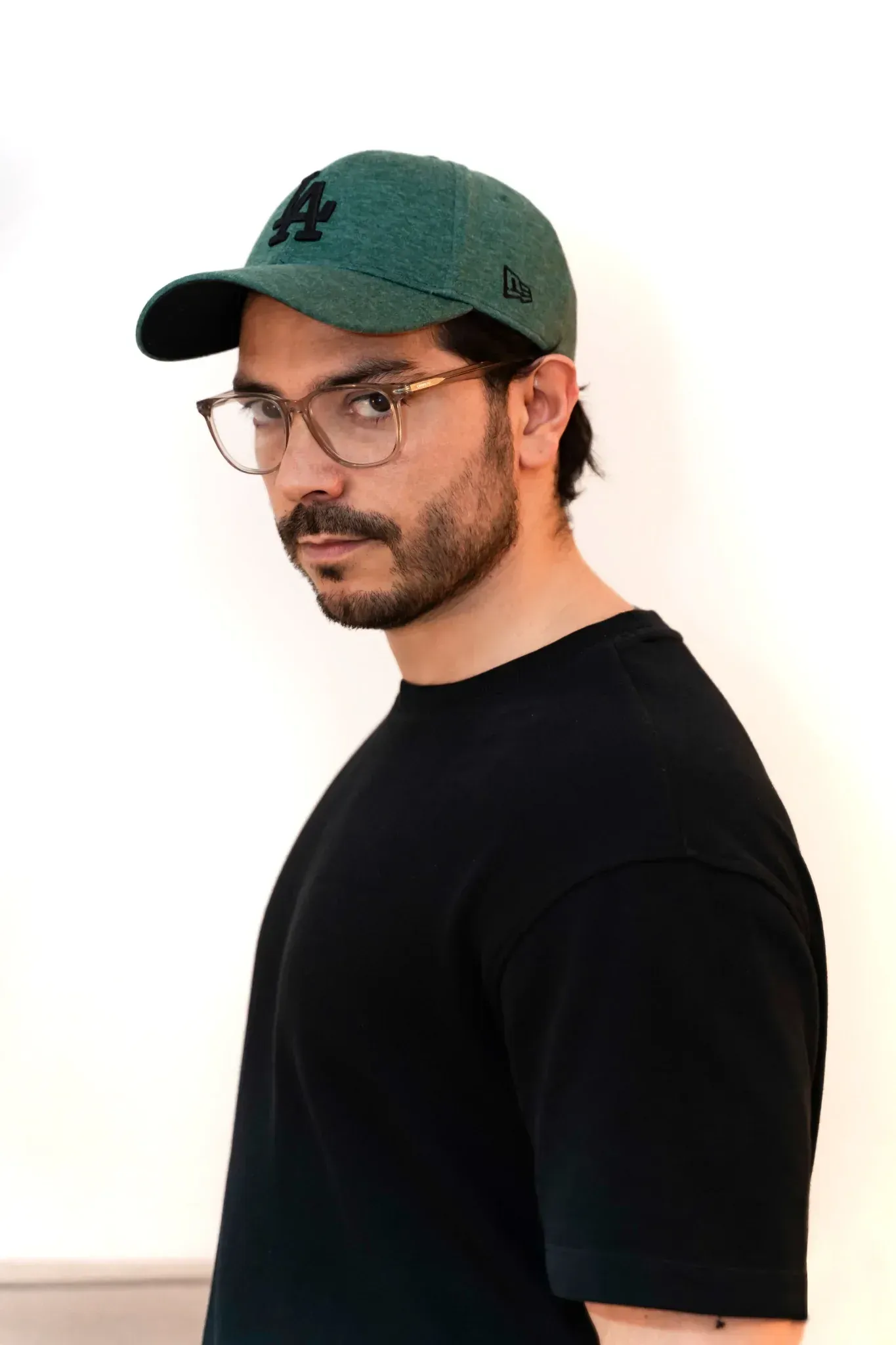 Hombre con gafas y gorra de béisbol verde, sonriendo. Camisa negra de cuello redondo sobre fondo blanco.