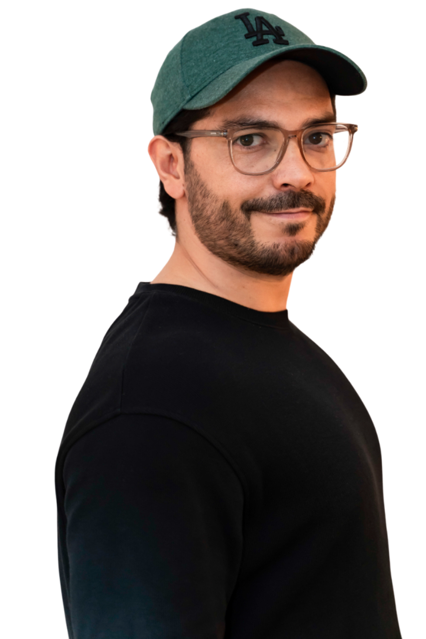 Hombre con gafas y gorra de béisbol verde, sonriendo. Camisa negra de cuello redondo sobre fondo blanco.