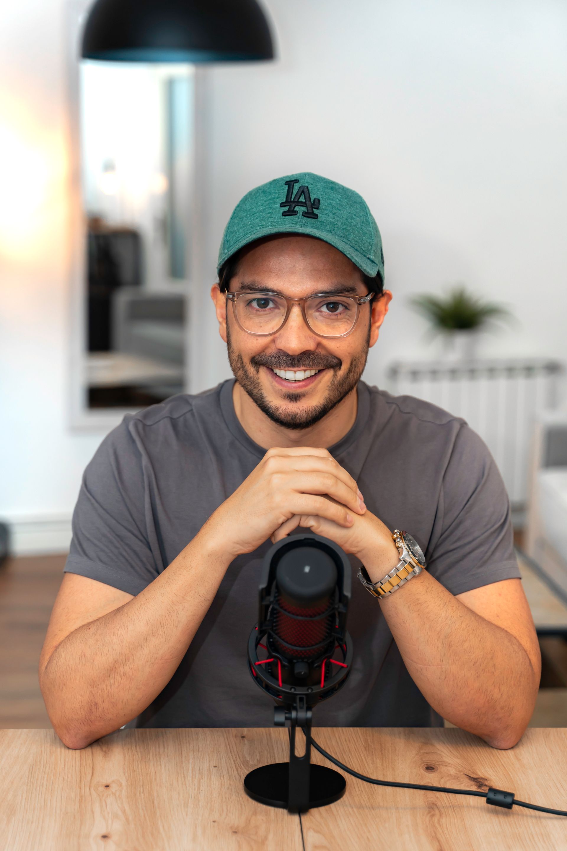Hombre con gafas y gorra verde, sonriendo ante un micrófono sobre un escritorio.