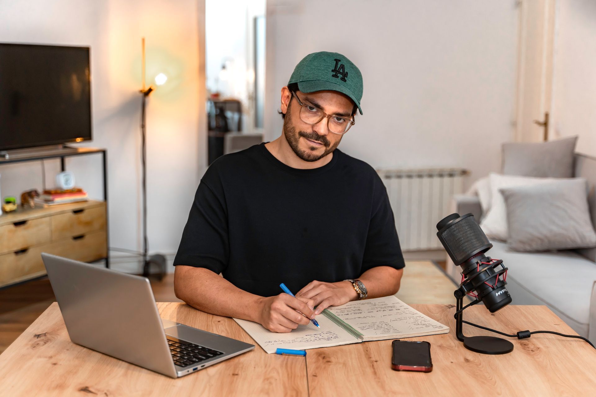 Un hombre con sombrero y gafas se sienta en un escritorio, escribiendo en un cuaderno, con una computadora portátil y un micrófono.