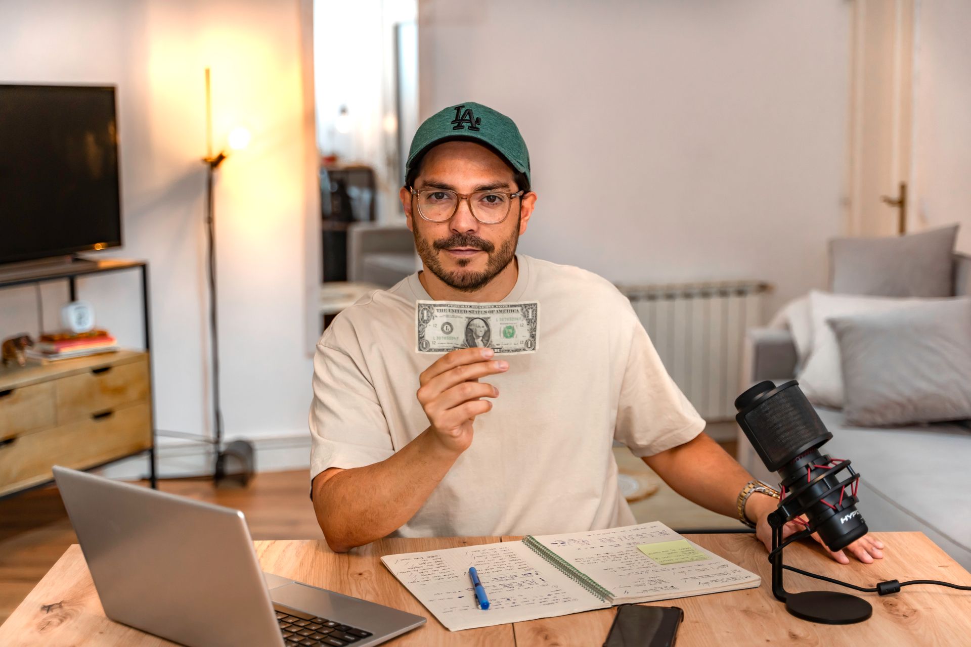Hombre sosteniendo un billete de un dólar, sentado en un escritorio con una computadora portátil, un micrófono, una libreta y una lámpara de fondo.