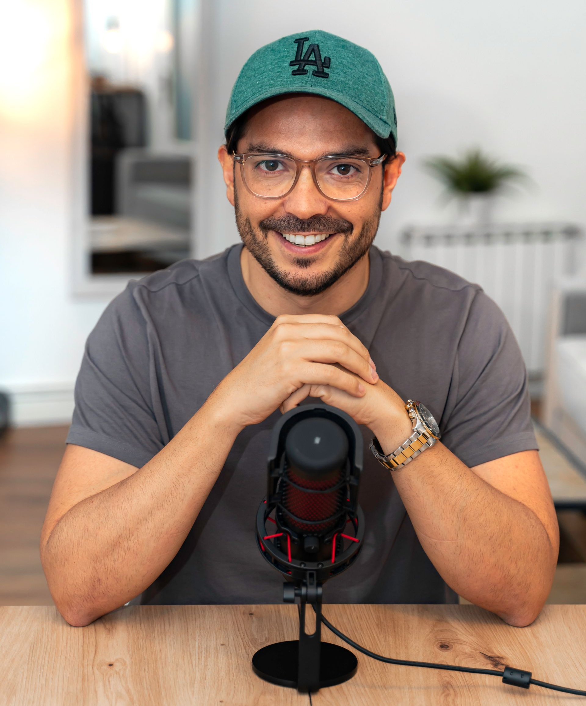 Hombre con gafas y gorra verde, sonriendo ante un micrófono sobre un escritorio.