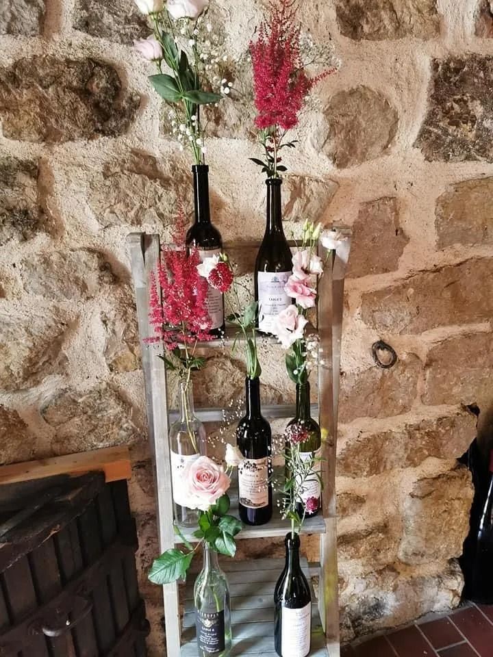 Des bouteilles de vin et des fleurs disposées sur une étagère rustique en bois, adossée à un mur de pierre.