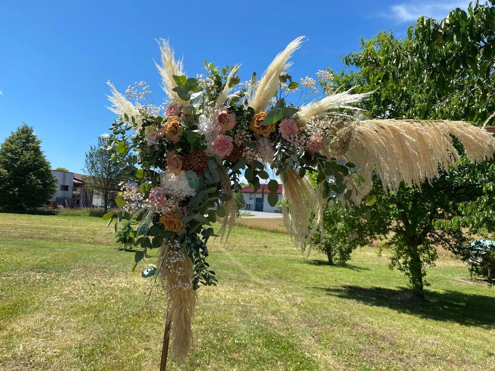Arche de mariage florale ornée d'herbes de la pampa et de fleurs roses, dans un cadre champêtre sous un ciel bleu.