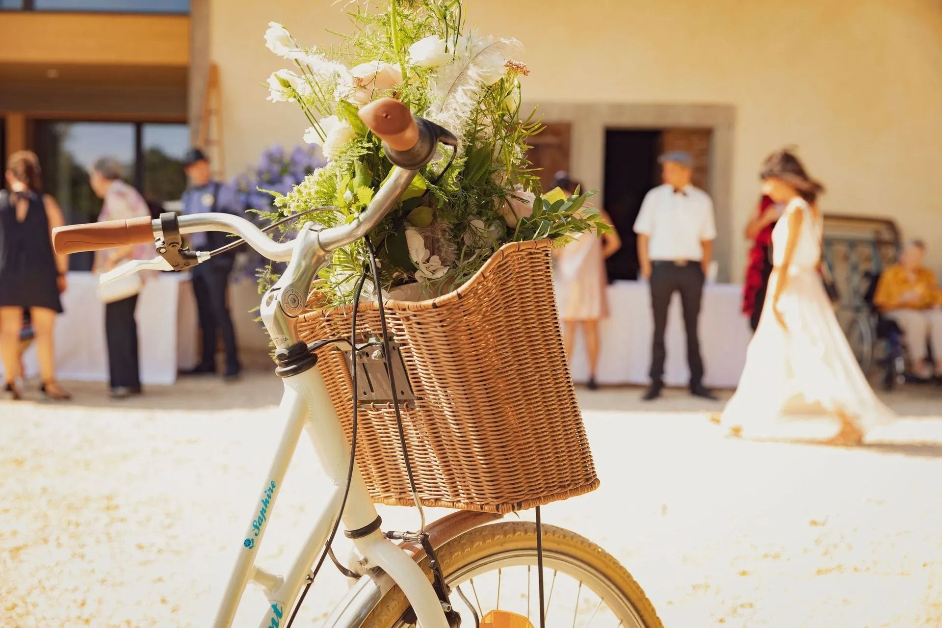 Un vélo transportant un panier de fleurs lors d'un mariage. La mariée et ses invités sont visibles en arrière-plan.