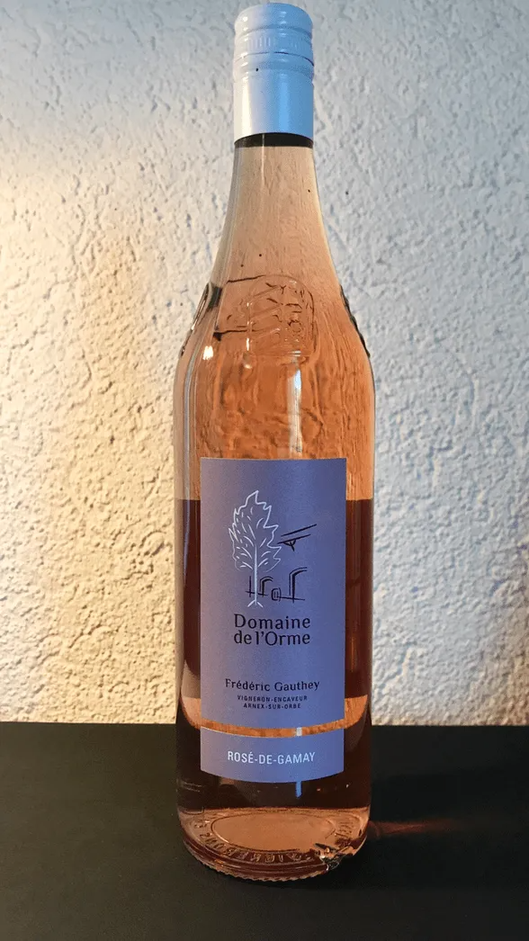 Bouteille de vin rosé à l'étiquette et au bouchon bleus, posée sur une surface sombre, devant un mur blanc texturé.