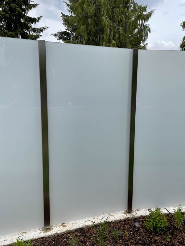 Ein weißer Glaszaun mit Edelstahlpfosten und Bäumen im Hintergrund.