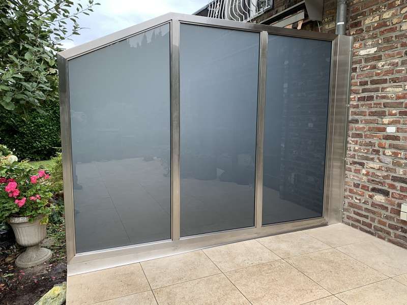 Auf einer Terrasse neben einer Backsteinmauer steht ein Edelstahlzaun mit grauem Glas.