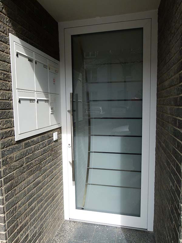 Neben einer Backsteinmauer steht eine weiße Tür mit Glaseinsatz.