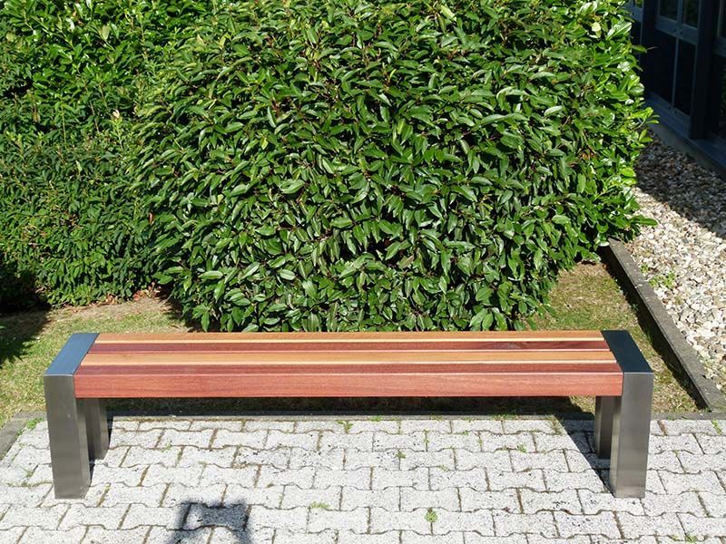 Eine Holzbank steht auf einem gepflasterten Gehweg vor einem Busch.