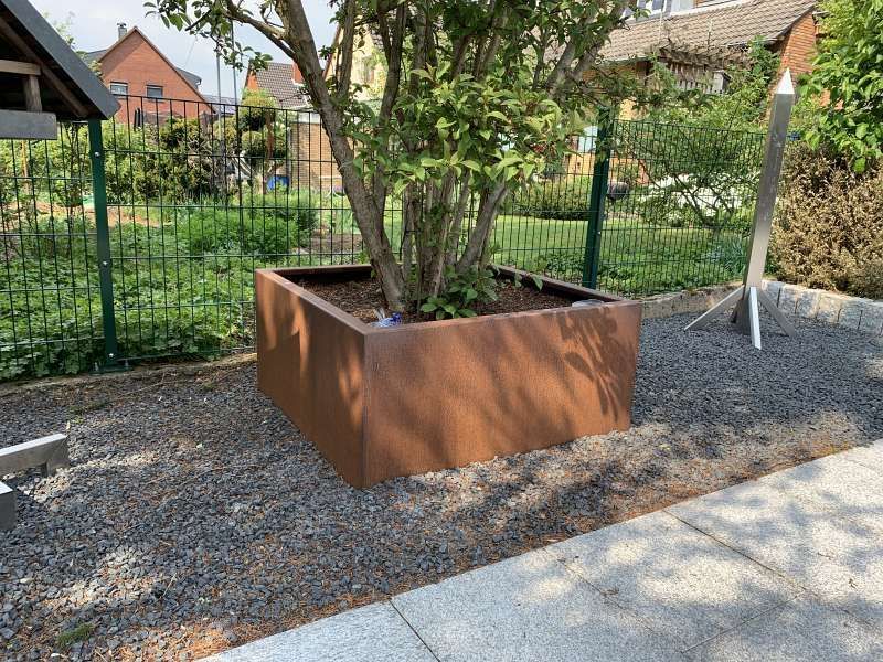 Ein großer brauner Blumenkasten mit einem Baum darin steht inmitten einer Kiesbereich.