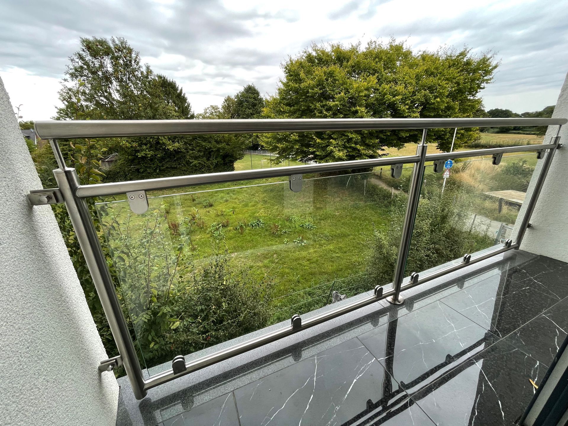 Balkon mit Glaspaneelen, Edelstahlgeländer und schwarzem Fliesenboden mit Blick auf eine Rasenfläche und Bäume.