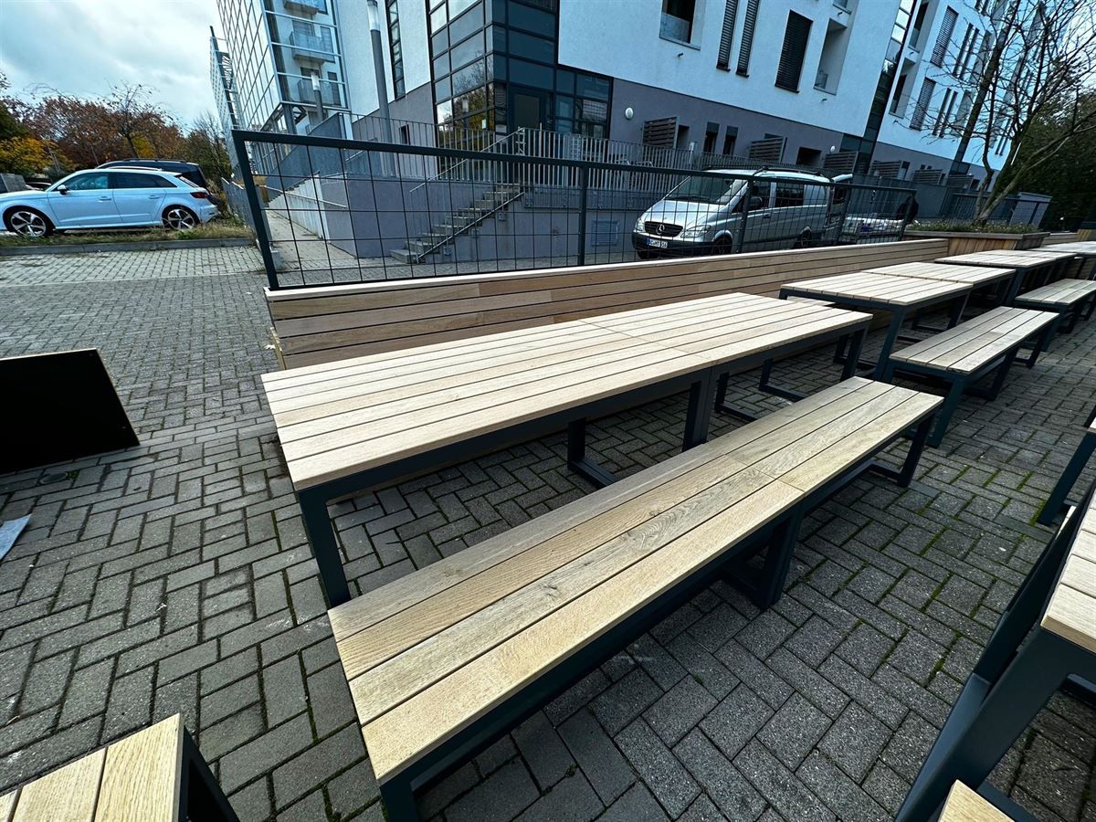 Eine Reihe von Holztischen und Bänken auf einem gepflasterten Gehweg vor einem Gebäude.