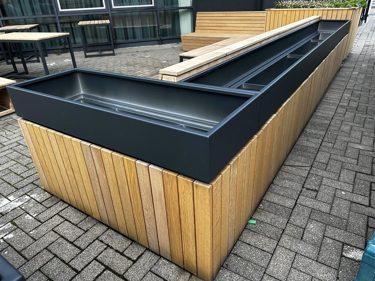 Auf einem gepflasterten Gehweg steht ein großer Blumenkasten aus Holz.