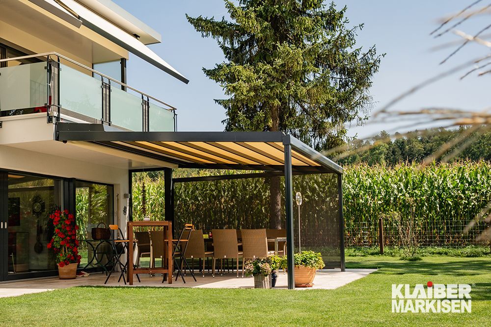 Eine Terrasse mit Pergola und Tisch und Stühlen vor einem Haus.