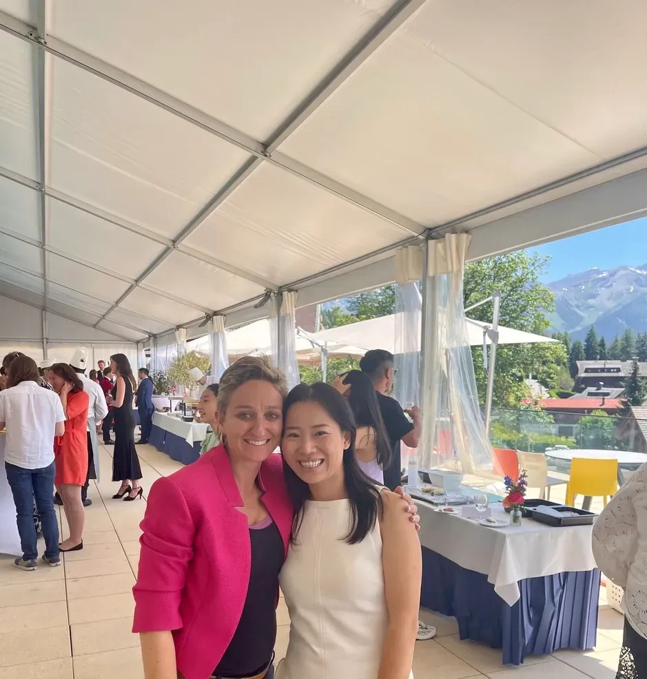 Deux femmes souriantes lors d'un événement en plein air sous une tente ; l'une en blazer rose, l'autre en robe blanche.
