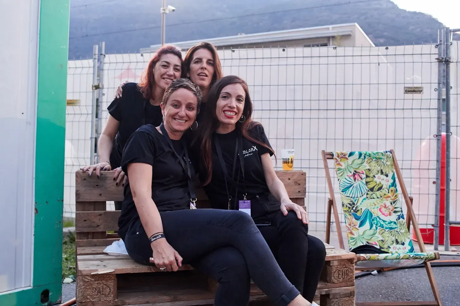 Quatre femmes en chemises noires sont assises sur des palettes en bois, souriantes, près d'une clôture.