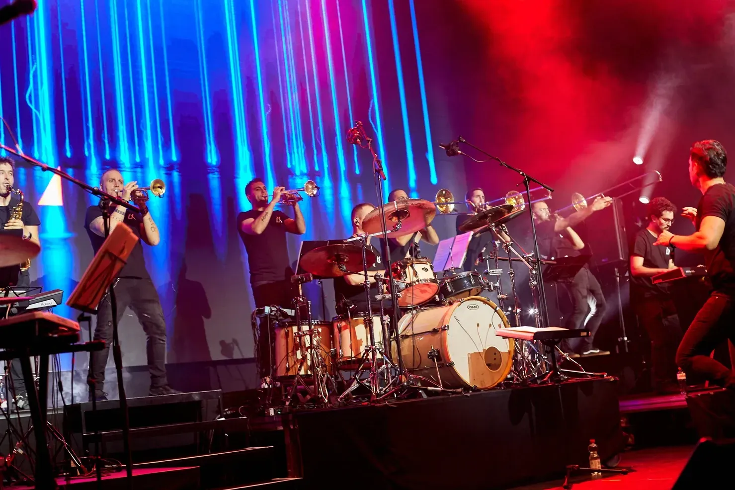 Groupe sur scène, avec cuivres, batterie et claviers, jouant sous des lumières de scène bleues et rouges.