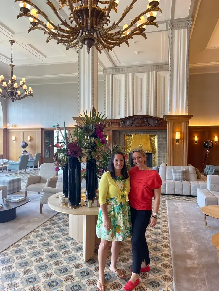 Deux femmes souriantes dans le hall d'un hôtel chic avec un grand lustre et des compositions florales.