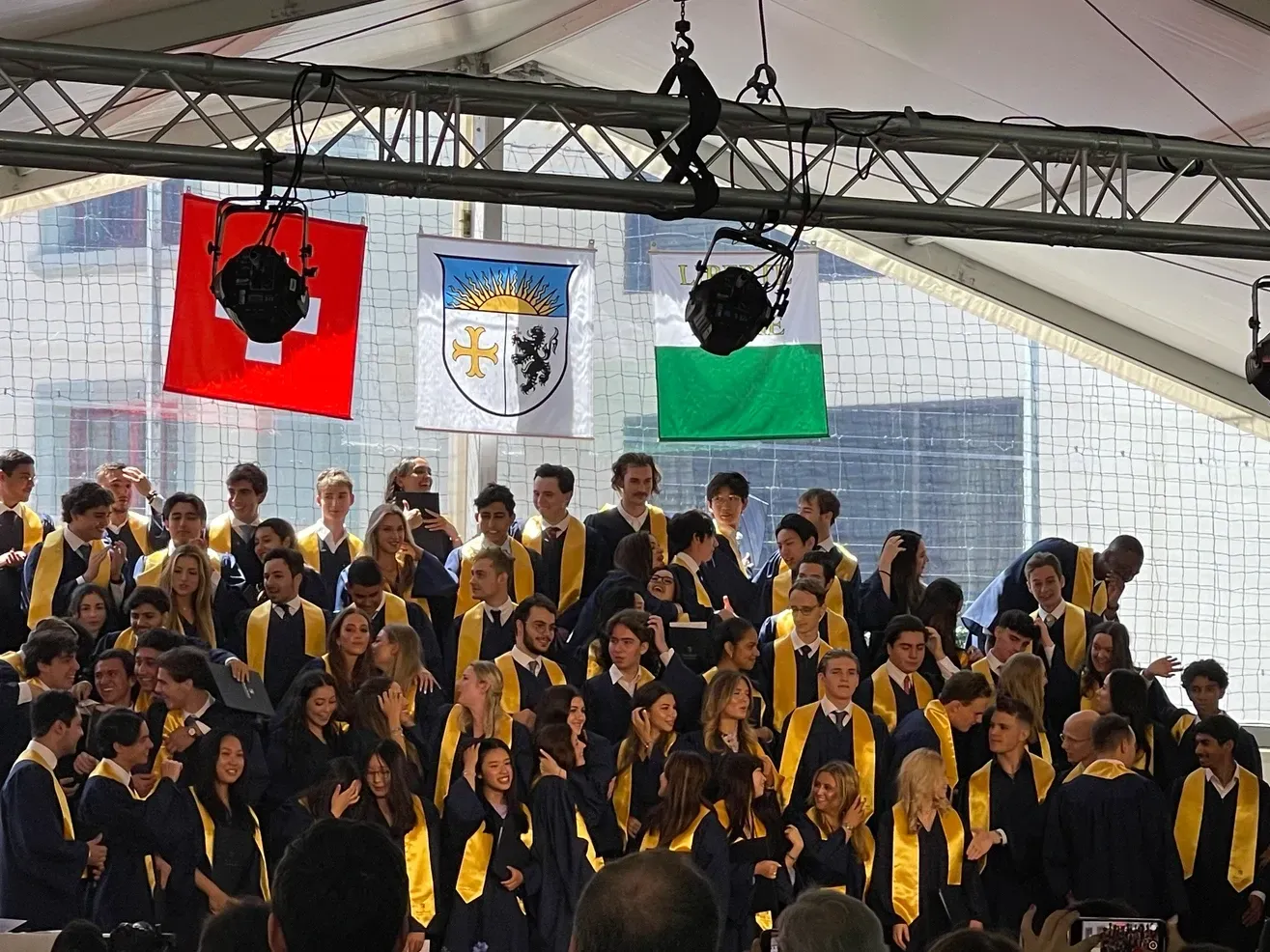 Cérémonie de remise des diplômes avec des étudiants en toges bleues, drapeaux suisses, écusson et vert.