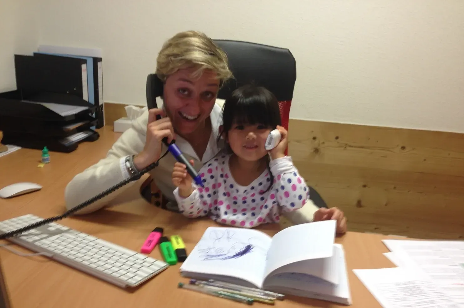 Femme et enfant à leur bureau, tous deux tenant leur téléphone. Une femme blonde sourit, un enfant dessine.