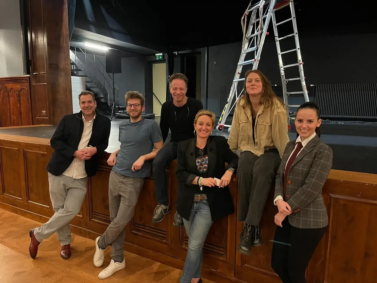 Six personnes posent nonchalamment sur une scène en bois, avec une échelle en arrière-plan.