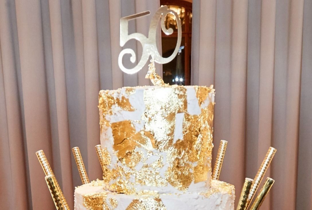 Gâteau blanc à trois étages, décoré de feuilles d'or et orné du chiffre « 50 ». Des pailles dorées entourent la base