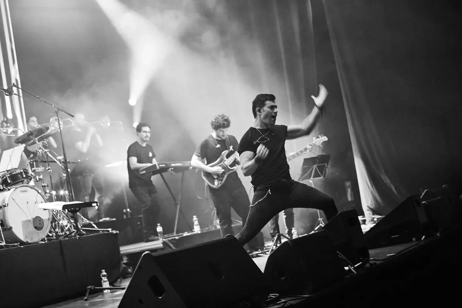 Groupe sur scène, chanteur le bras levé, devant ses camarades. Photo en noir et blanc.