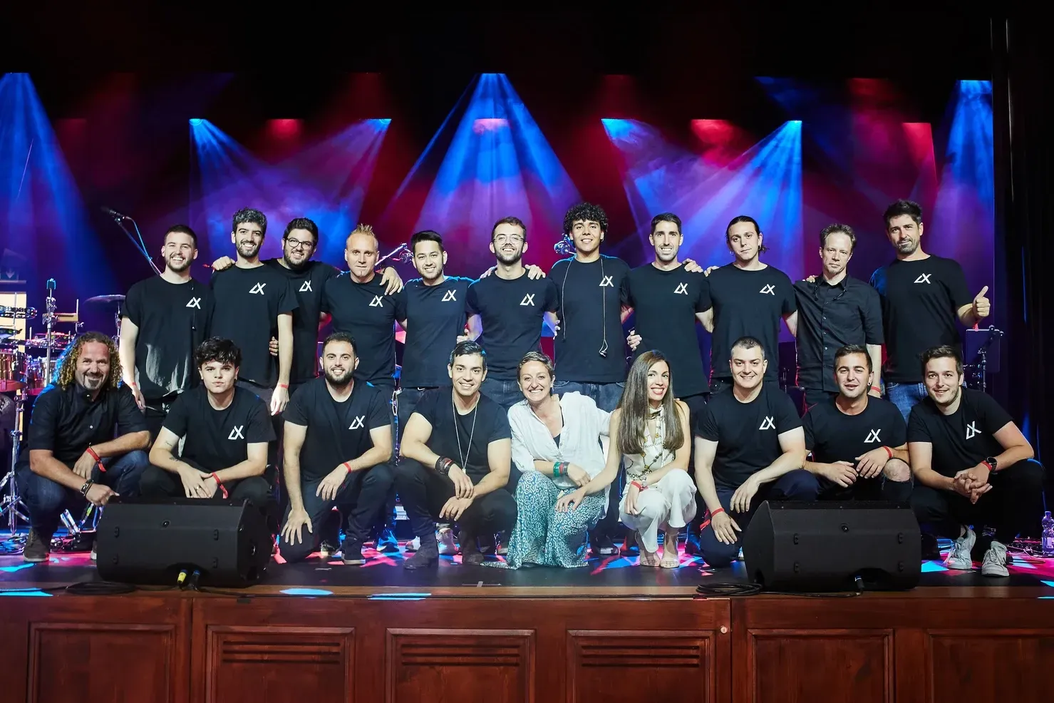 Groupe de musiciens sur scène, souriant à la caméra, sous des lumières bleues et rouges.