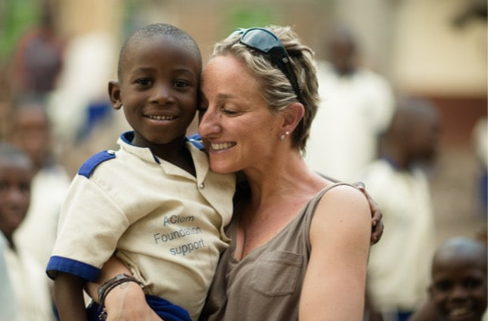 Une femme embrasse un enfant noir souriant en uniforme scolaire ; d'autres enfants en arrière-plan.