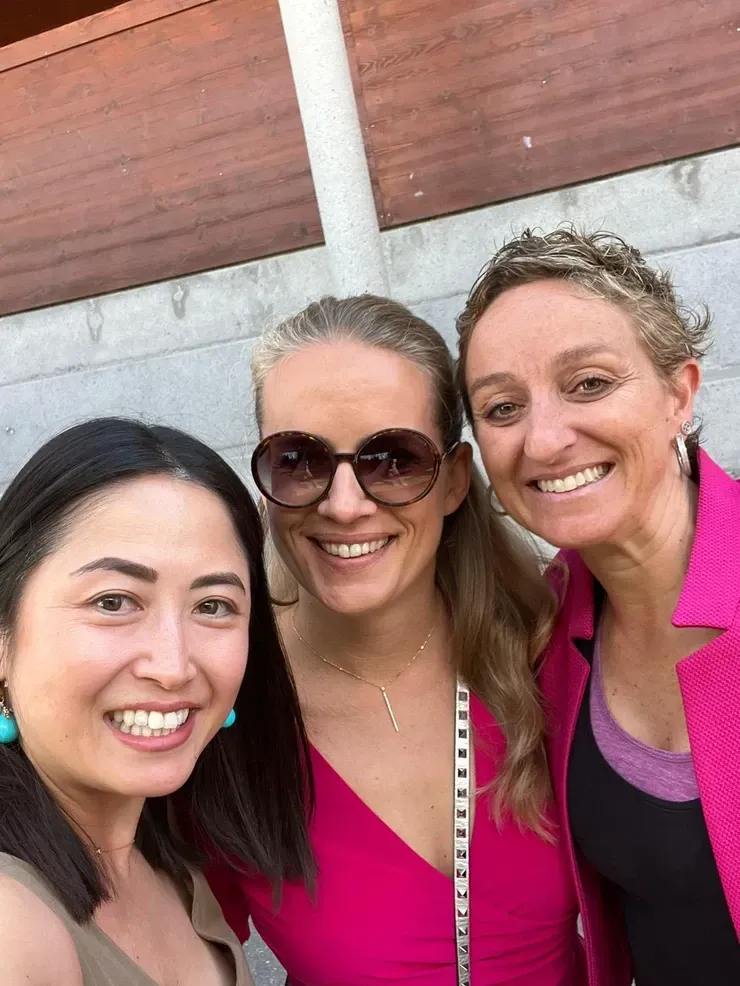 Trois femmes souriantes prennent un selfie en extérieur. L'une porte un blazer rose, l'autre une robe rose