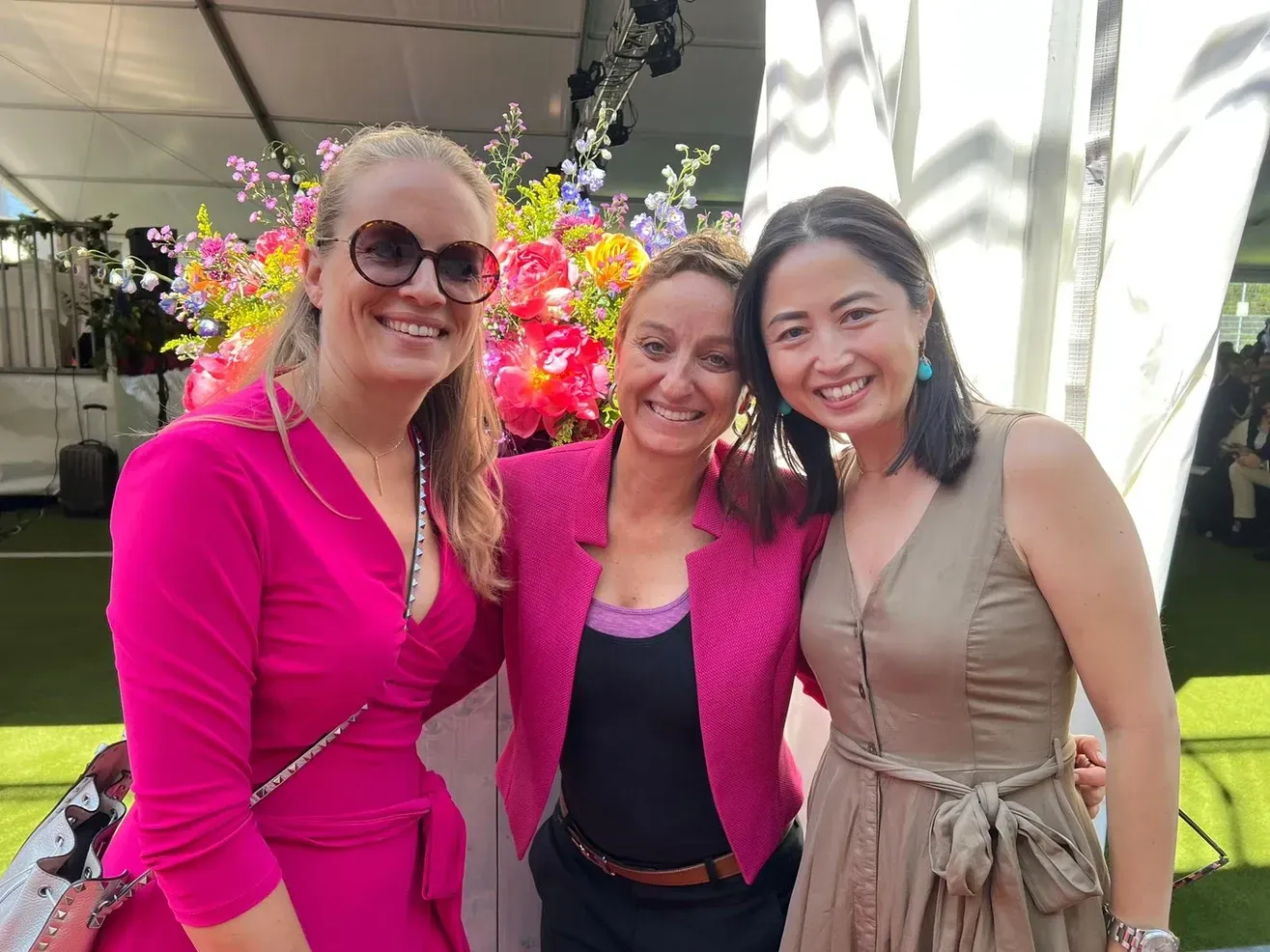 Trois femmes souriantes, posant ensemble à l'extérieur près des fleurs, l'une portant une tenue rose vif