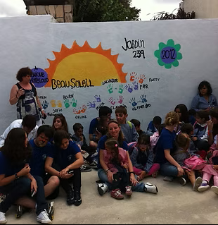 Enfants et adultes posent devant une fresque murale avec un soleil, des empreintes de mains et des noms