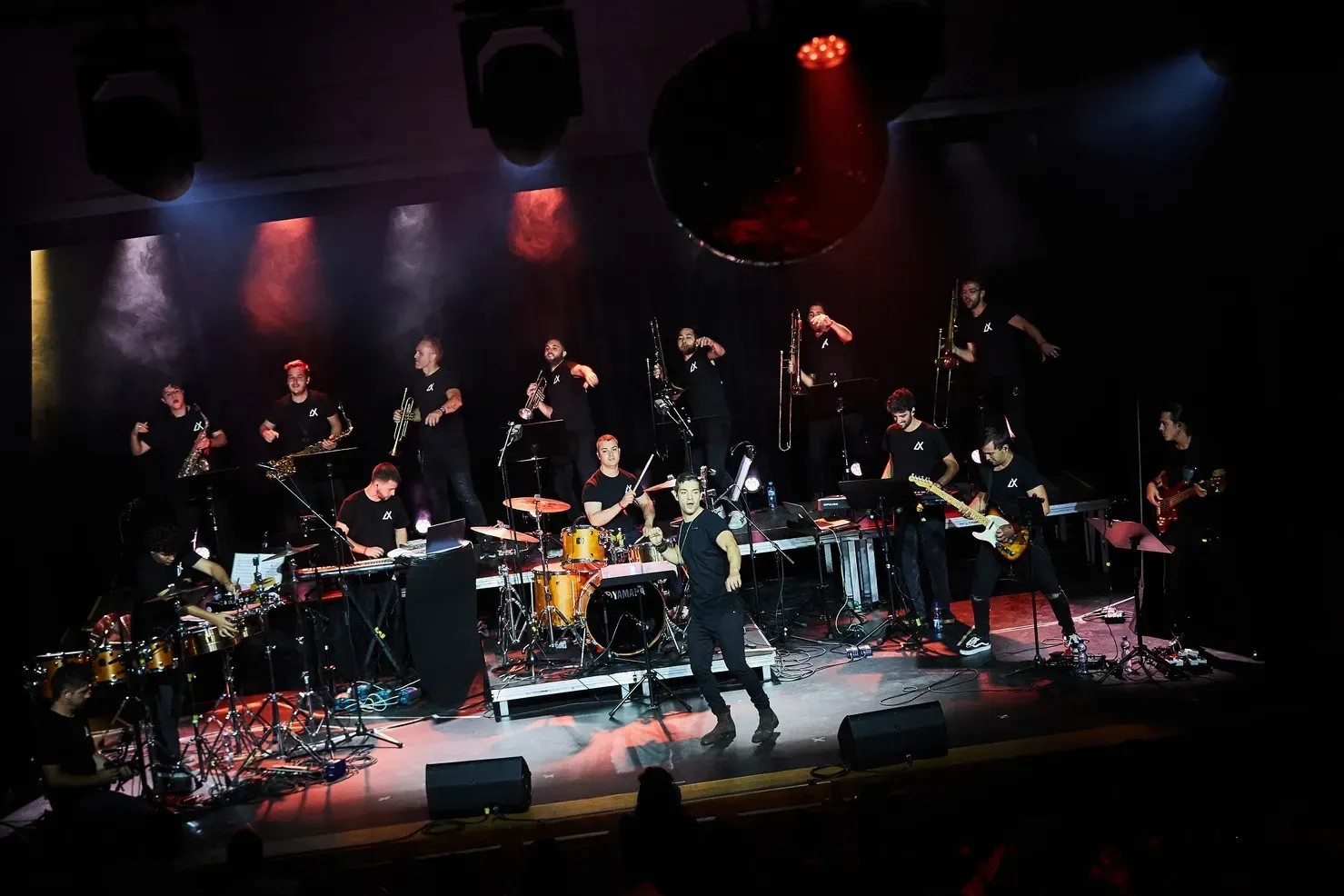 Un grand groupe sur scène avec des instruments, jouant sous des lumières rouges et bleues. Les musiciens sont vêtus de noir.