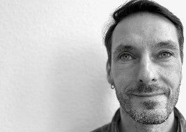 Mann mit kurzem Haar und Bartstoppeln, der mit einem leichten Lächeln in die Kamera blickt; er trägt einen kleinen Ohrring.