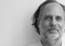 Ein Mann mit Bart und welligem Haar lächelt und lehnt an einer weißen Wand.