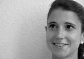 Eine Frau mit dunklen Haaren lächelt und trägt einen Ohrstecker vor einer strukturierten weißen Wand.