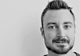 Mann mit kurzem, dunklem Haar und Bart, der leicht lächelt, vor einem schlichten Hintergrund.