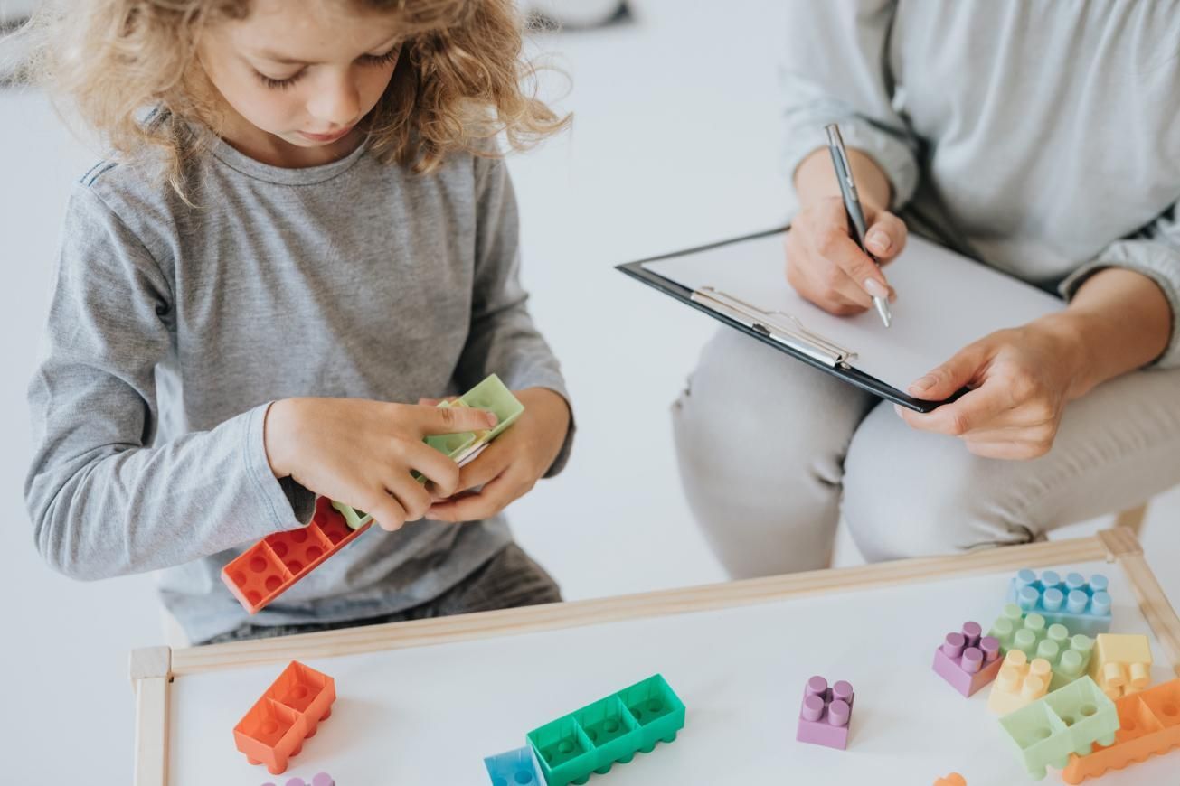 Un niño juega con bloques de construcción de colores mientras un adulto se sienta cerca y toma notas en un portapapeles.