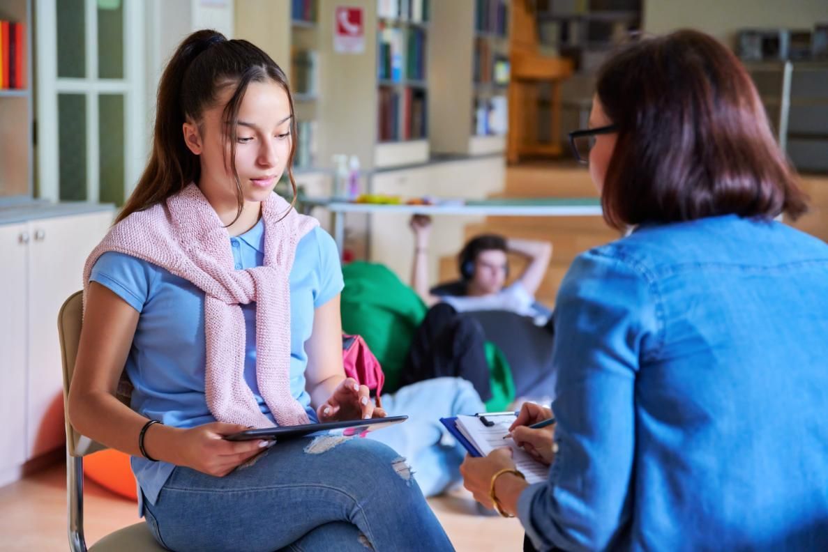 Un estudiante interactúa con un profesional en un entorno de biblioteca, mientras otra persona descansa al fondo.