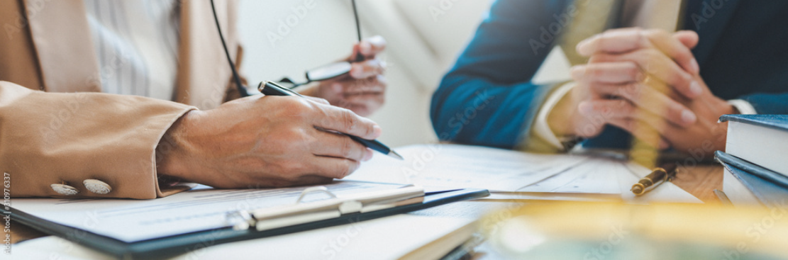 Zwei Personen prüfen ein Dokument; eine schreibt auf einem Klemmbrett, die andere zeigt mit einem Stift darauf.