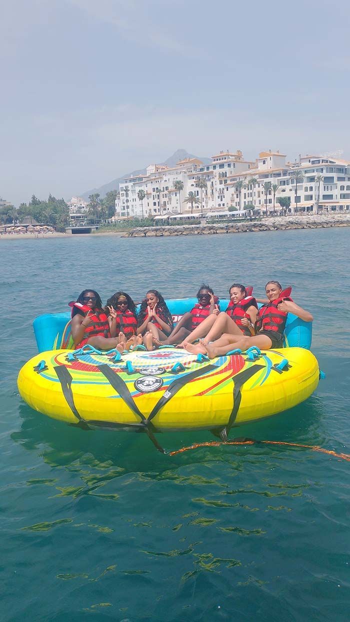 Seis personas en una balsa inflable amarilla, con chalecos salvavidas, en el océano. Edificios blancos al fondo.