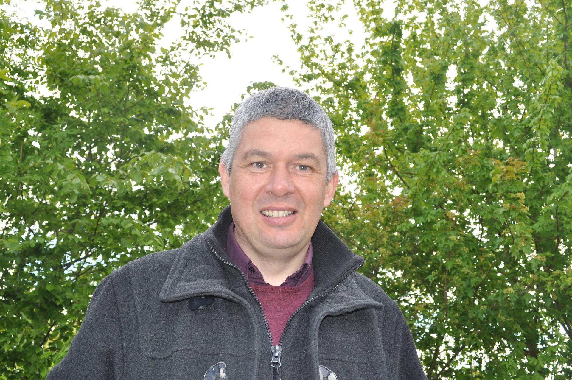 Un homme aux cheveux grisonnants sourit en extérieur, vêtu d'une veste sombre sur une chemise bordeaux.