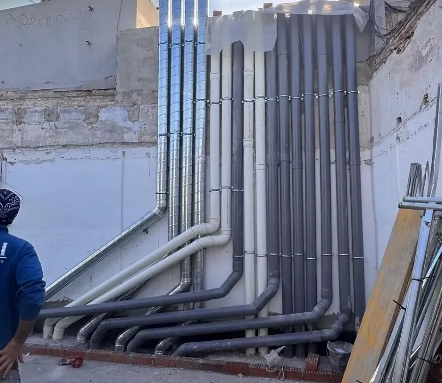 Una hilera de tuberías verticales de metal, PVC blanco y gris oscuro, montadas contra una pared de construcción sin terminar.