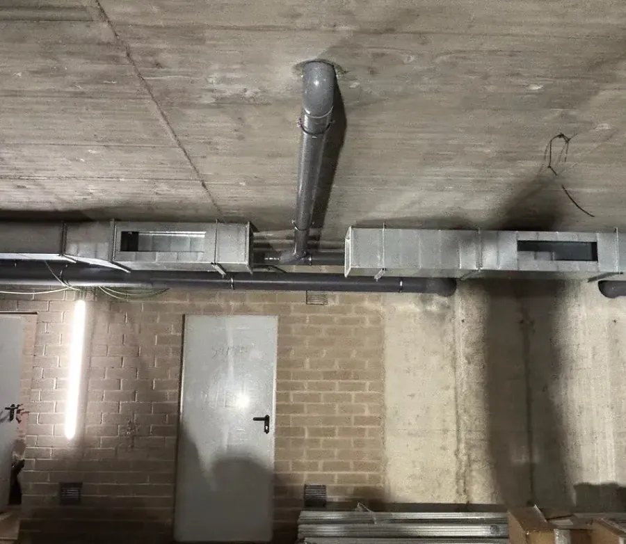 conductos de ventilación metálicos y una tubería gris suspendida de un techo de hormigón sobre una puerta.