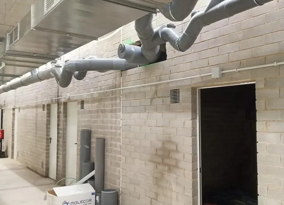 Los conductos de ventilación a la vista recorren la parte superior de un pasillo con puertas cerradas y una puerta abierta.