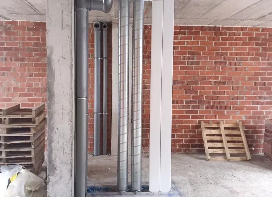 Pared de ladrillo visto en una obra en construcción, con tuberías verticales grises, conductos de ventilación metálicos.