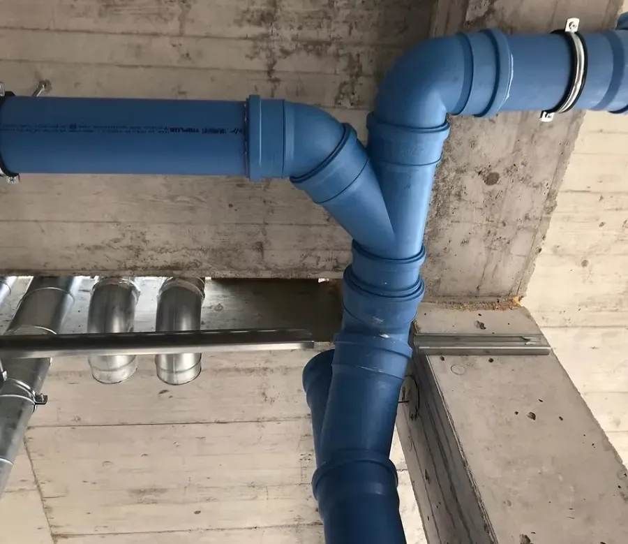 Tuberías de desagüe conectadas mediante una pieza en forma de Y, suspendidas de un techo junto a conductos metálicos.