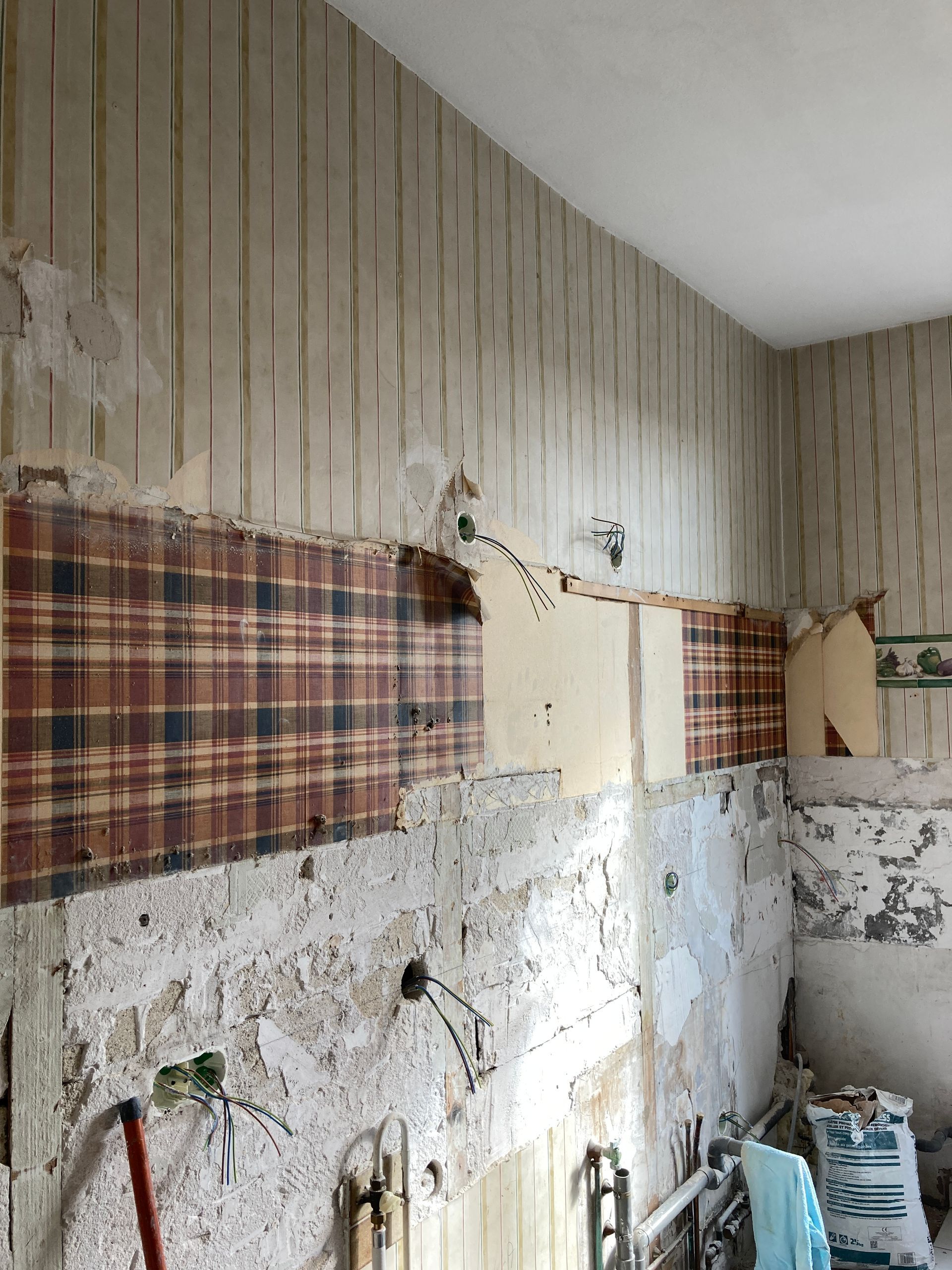 Mur dont le papier peint partiellement décollé laisse apparaître un motif à carreaux et des plaques de plâtre apparentes.